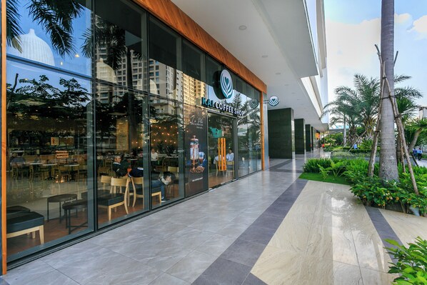 A modern coffee shop with large glass windows, outdoor seating, and a view of palm trees and buildings. - Refined Saigon - Rivergate Apartment (Ho Chi Minh City)