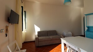 Ein Zimmer mit einer Couch, einem weißen Tisch, einer blauen Hängelampe und einem an der Wand befestigten Fernseher.