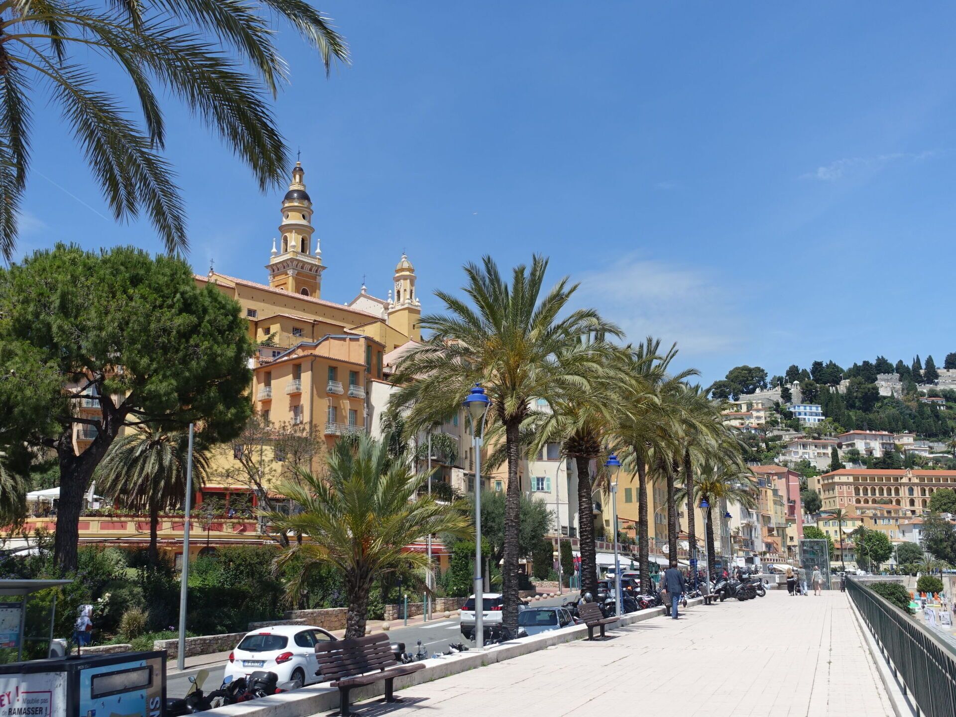 A coastal cityscape with a prominent church, palm trees, and a pedestrian walkway.