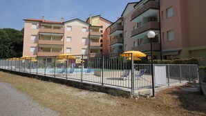 A multi-story residential building with balconies, a swimming pool area with sun loungers, and a fenced garden. - Comfy flat in Rosolina with shared pool (Rosolina Mare)