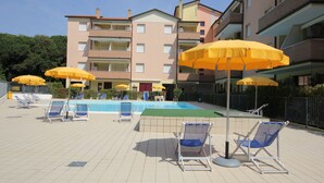 A pool area with lounge chairs and yellow umbrellas, surrounded by apartment buildings. - Comfy flat in Rosolina with shared pool (Rosolina Mare)