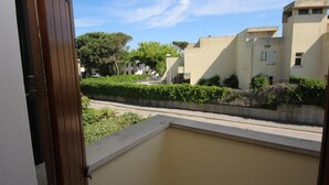 Uitzicht vanaf een balkon met een betonnen rand en een deur, op een straat met gebouwen en groen.