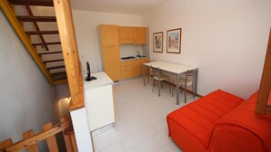Een compacte keuken met een eettafel en stoelen, een kleine koelkast, en een trap met houten leuningen.