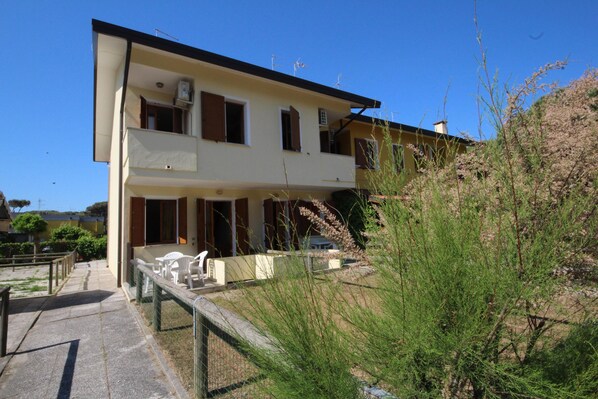 A two-story building with a balcony, a small garden area with chairs, and a clear blue sky.