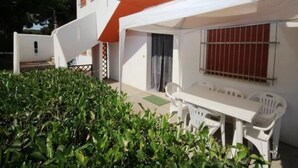 A white building with a red roof, a patio with a table and chairs, and a hedge in the foreground. - Comfortable beachside apartment in Rosolina (Rosolina Mare)