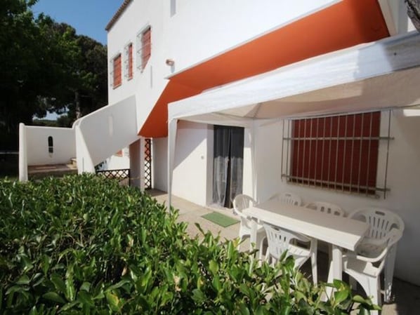 A white building with a red roof, a patio with a table and chairs, and a hedge in the foreground.