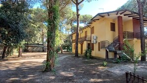 A yellow building with a balcony, surrounded by trees and a stone pathway. - Comfortable apartment in lovely Rosolina Mare (Rosolina Mare)