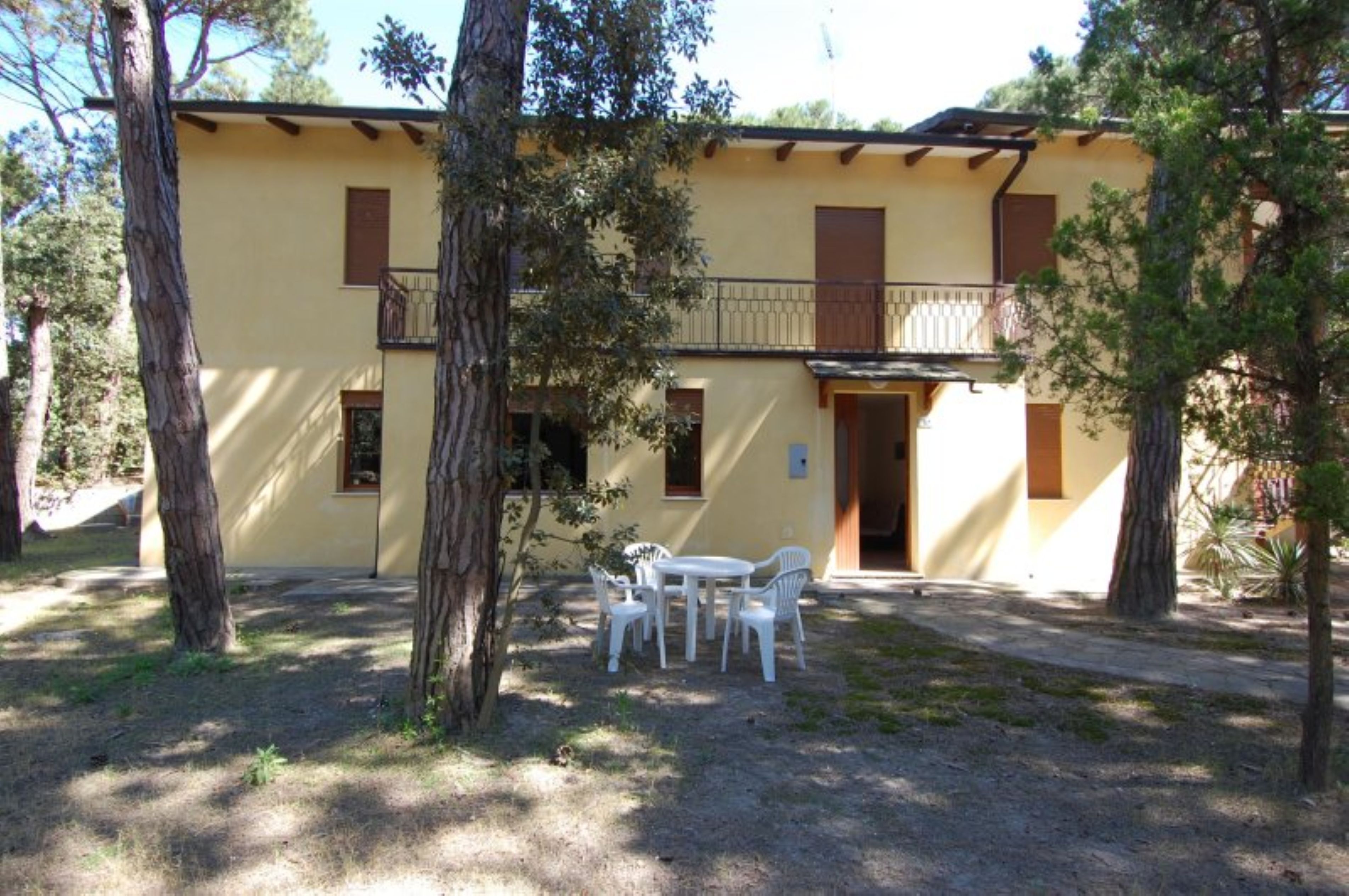 A two-story building with a balcony, a patio with a table and chairs, and trees in the foreground.