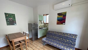 A compact living space with a wooden dining table and chairs, a sofa with a patterned cover, a kitchen area with a refrigerator, and a window with a view. - Accommodation with shared garden close to the sea (Rosolina Mare)