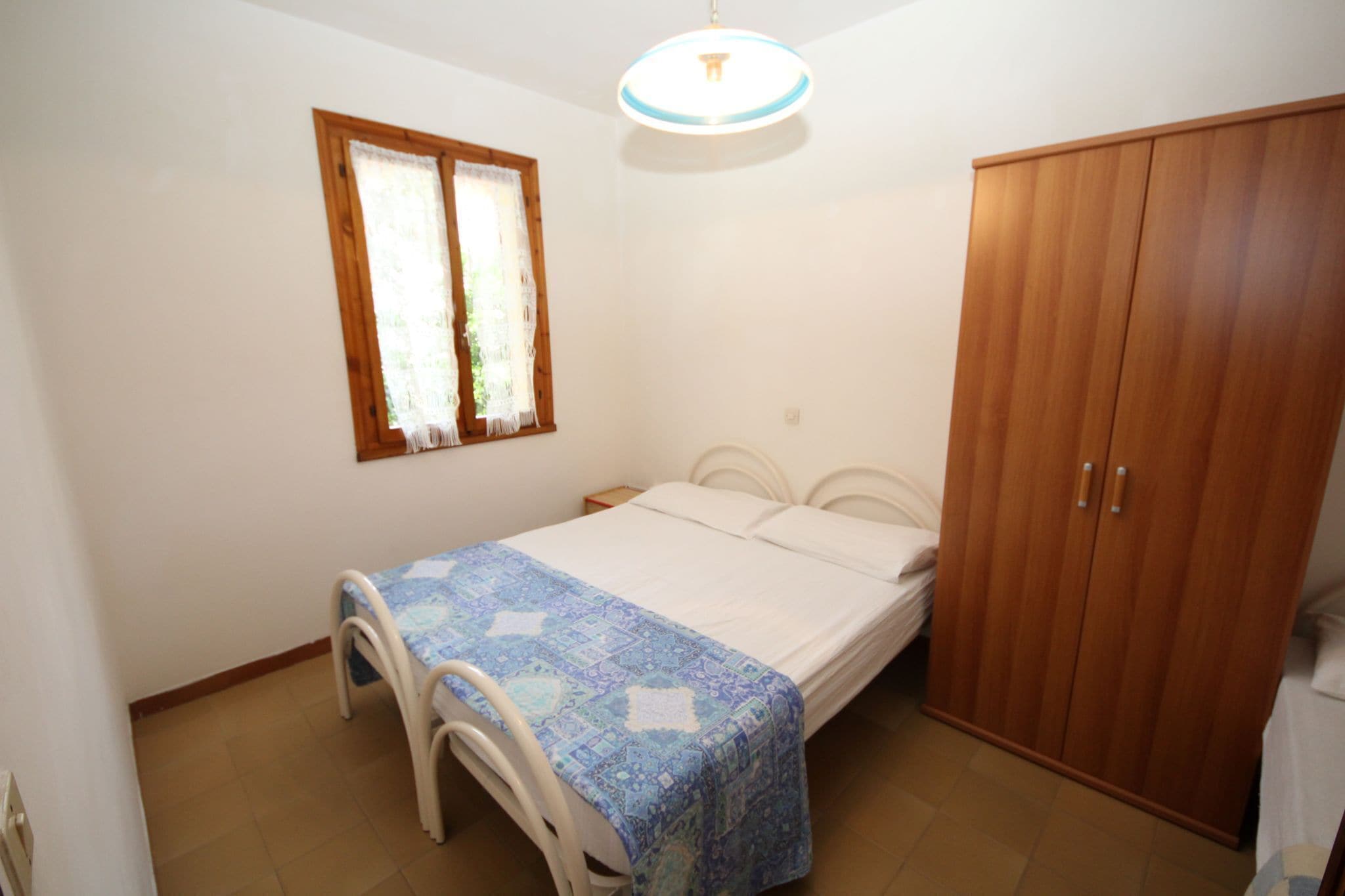 A bedroom with a bed, a wooden wardrobe, a window with curtains, and a ceiling light fixture.