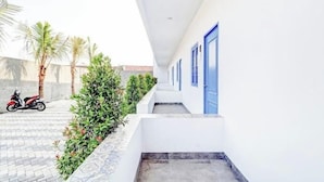 A corridor with white walls and blue doors, leading to a covered area with a tiled floor and a row of palm trees.
