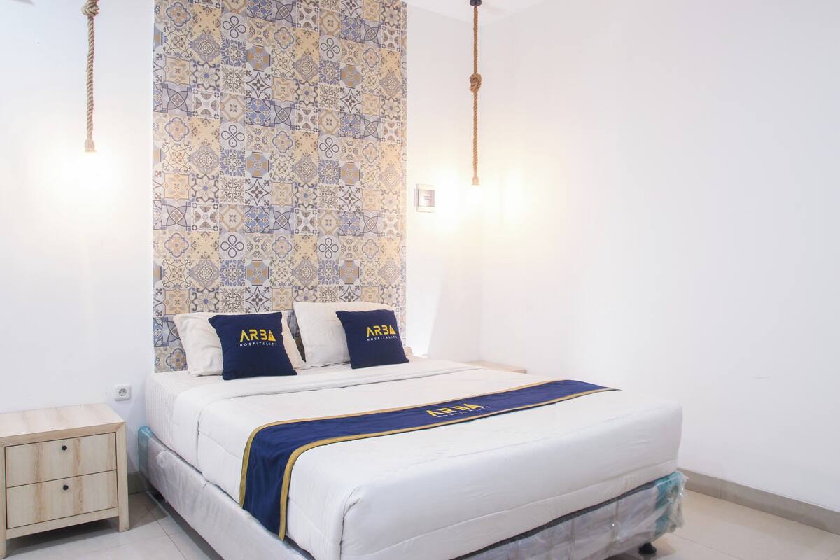 Un lit bien fait, recouvert d’une couvre-lit bleue et dorée et agrémenté d’oreillers, un mur orné d’un motif et une petite table de chevet en bois.