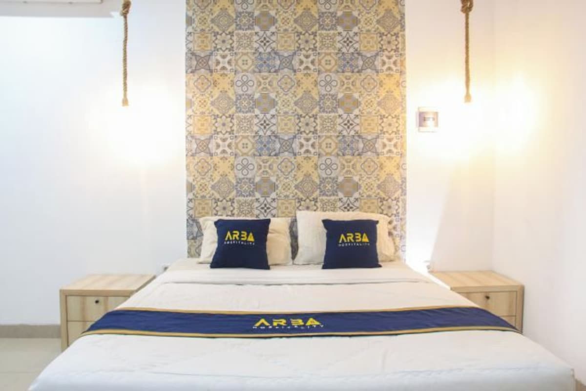 A hotel room with a bed, two nightstands, patterned wallpaper, and a bedspread with the logo "AR3A".