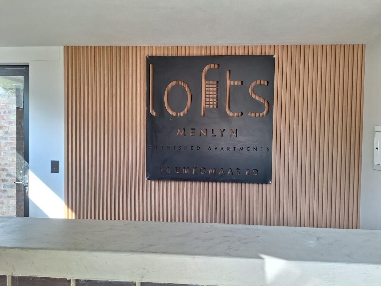 A sign for 'Lofts' with 'Menlyn Furnished Apartments' and '76 Umkomaas' written on it, mounted on a wooden wall.