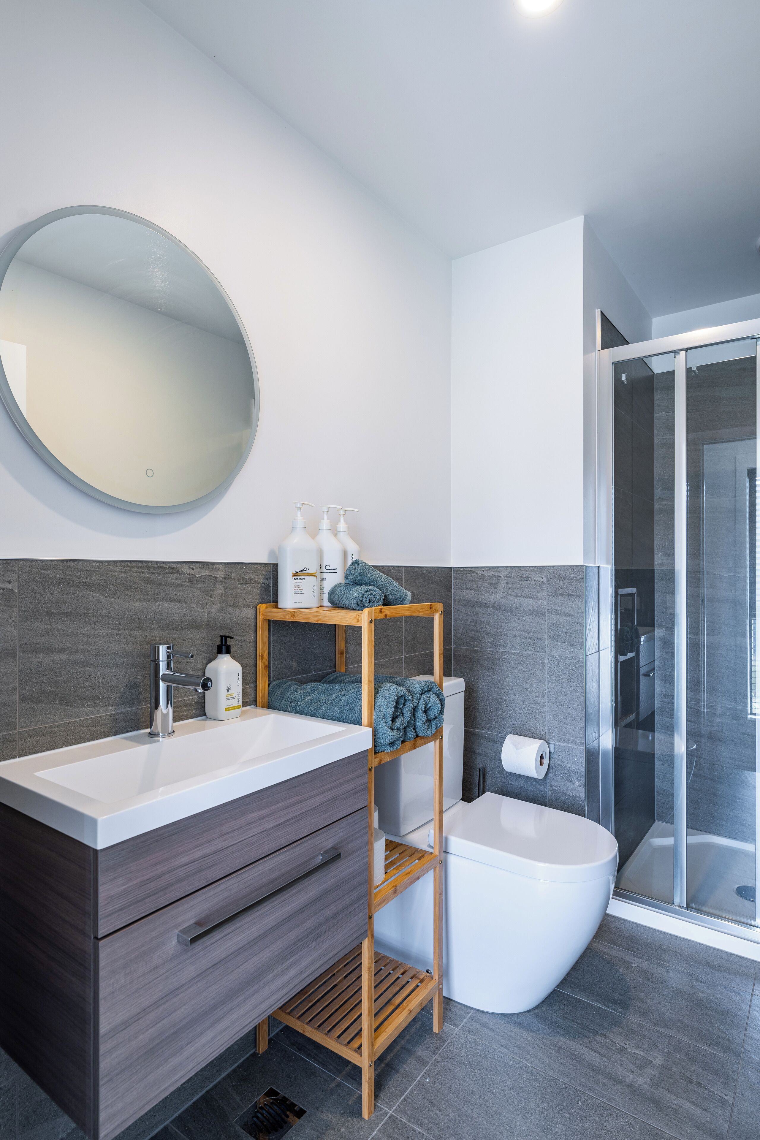 Une salle de bain moderne avec un lavabo blanc, un meuble-lavabo en bois foncé, un miroir rond et une cabine de douche en verre.
