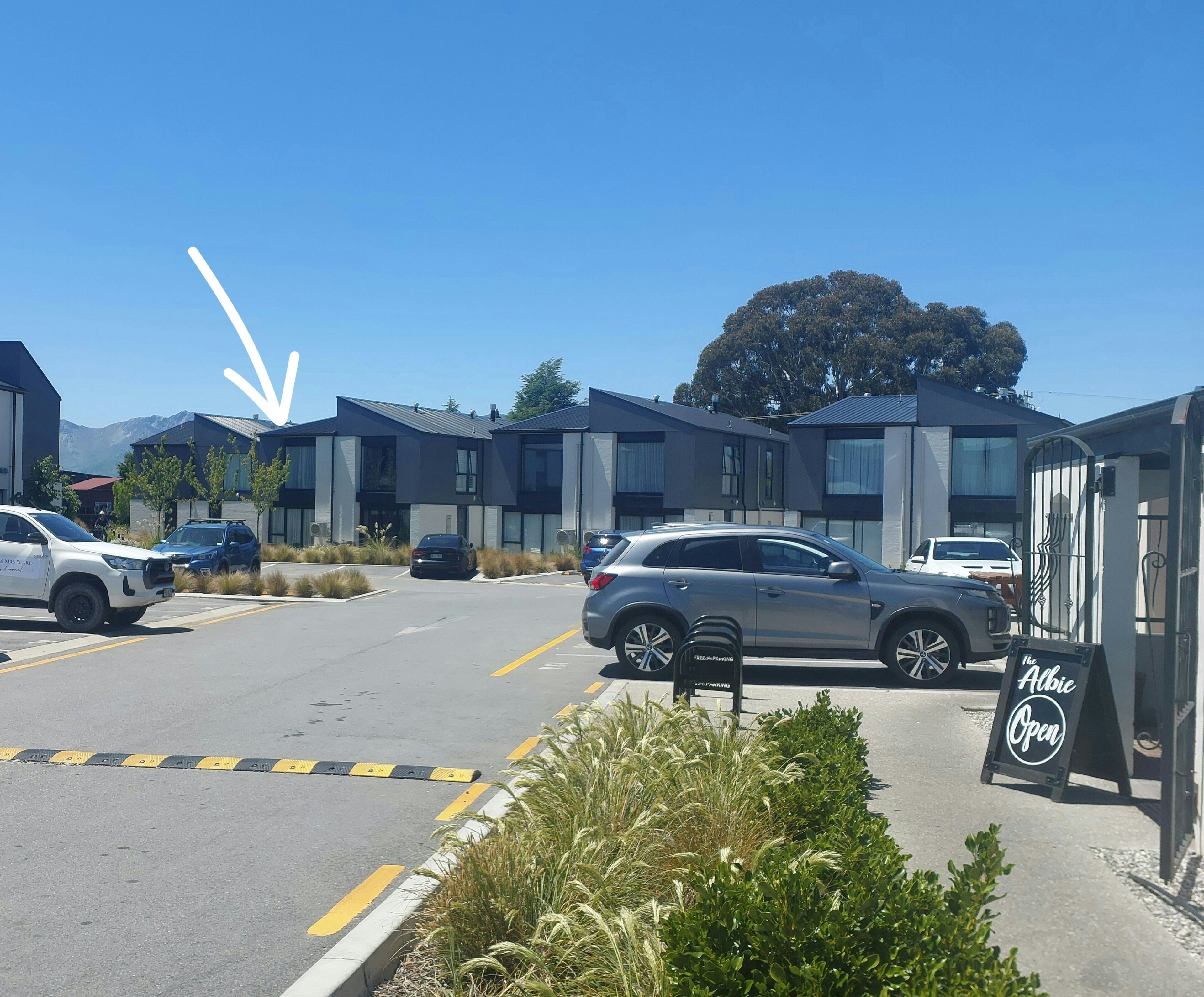 A parking lot with modern buildings, a sign for 'The Albie Open', and a mountain in the background.