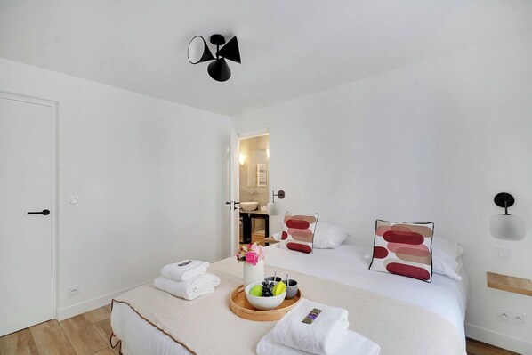 A hotel room with a bed, a bedside table with a bowl of fruit and a vase of flowers, a mirror, and a wall-mounted light fixture.