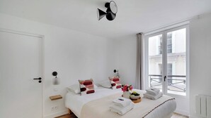 A modern bedroom with a large bed, a bedside table, a small shelf, a wall-mounted light, and a window with a view of buildings. - Chic apartment - 2BR/6P - Saint-Ambroise (Paris)
