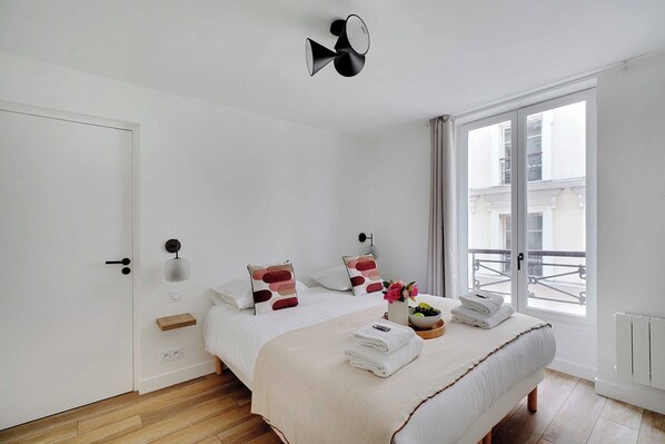 Un dormitorio moderno con una cama grande, una mesita de noche, un estante pequeño, una lámpara de pared y una ventana con vistas a edificios.
