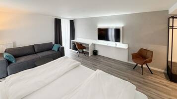 A modern hotel room with a large bed, a sofa, a desk with a TV, and a chair.