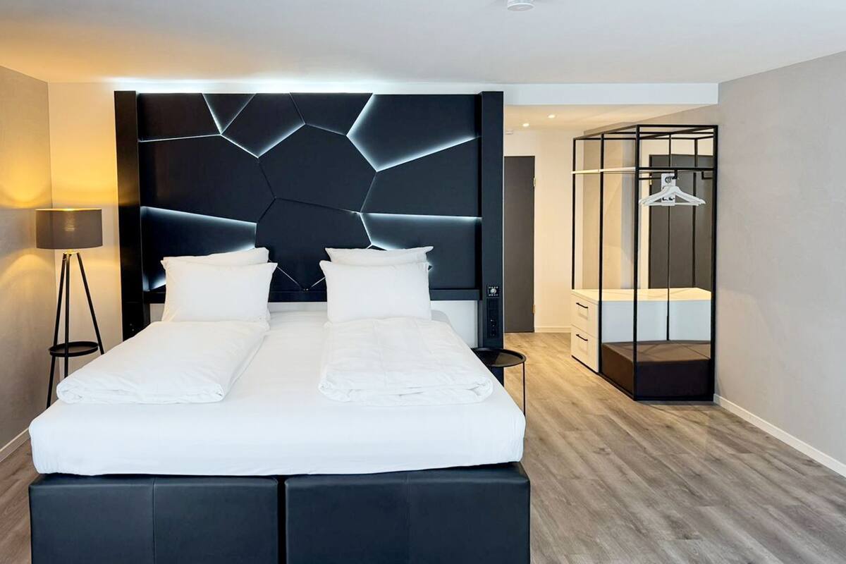 Una habitación de hotel moderna con una cama grande, cabecera negra con motivos geométricos, una mesita de noche, una lámpara de pie y un vestidor con perchero.