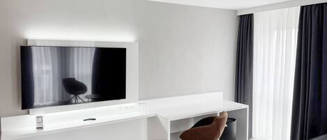 Ett modernt hotellrum med ett vitt skrivbord, en platt-TV, en brun stol och ett stort fönster med mörka gardiner.