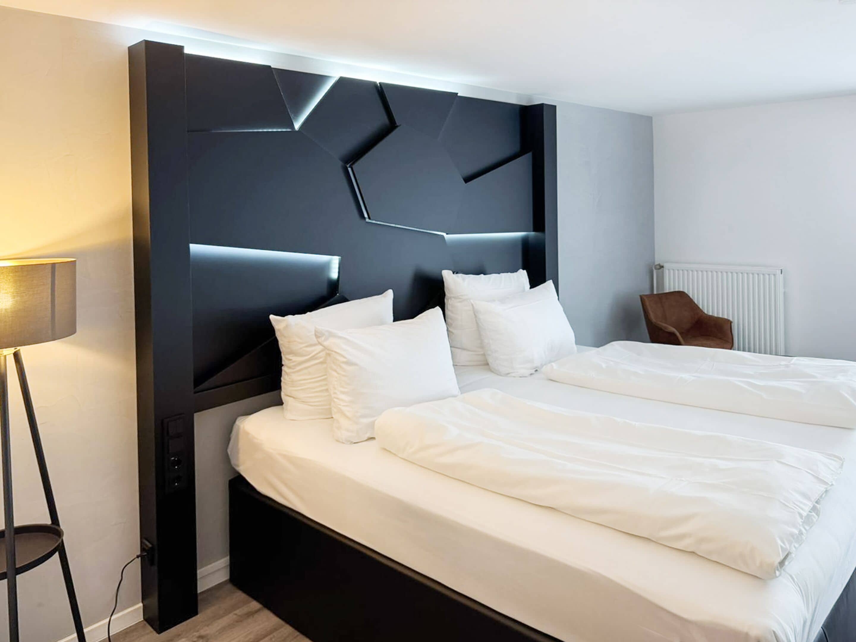A modern hotel room with a large bed, a dark headboard, and a side table with a lamp.