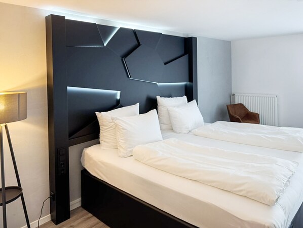 A modern hotel room with a large bed, a dark headboard, and a side table with a lamp. - DORMERO Hotel Weingarten (Weingarten)