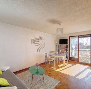 A living room with a sofa, a dining table with chairs, a small glass table with a vase, and a TV on a stand.