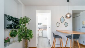 Una cocina moderna con armarios blancos, un microondas y un refrigerador pequeño. Hay una zona de comedor con una mesa de madera y dos sillas negras. Una planta grande en maceta está en primer plano, y un espejo cuelga en la pared.