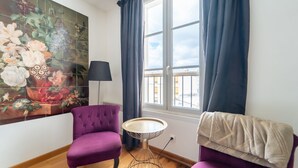 Una habitación con un gran mural floral, dos sillones morados, una pequeña mesa redonda, una lámpara y vistas de un edificio a través de una ventana con cortinas oscuras.