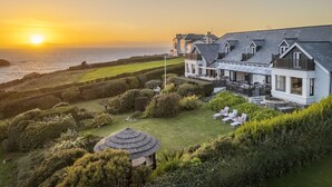 A large, modern house with a well-maintained garden and a view of the ocean at sunset. - Henscath House's characterful and spacious design perfectly caters for family and friends (Helston)