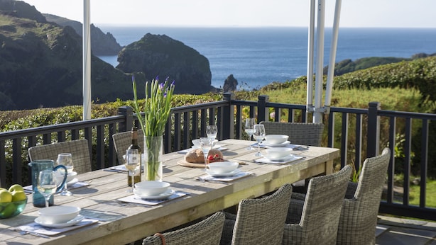 A wooden table set for dining with white plates, bowls, and glasses, overlooking a coastal landscape with cliffs and the sea. - Henscath House's characterful and spacious design perfectly caters for family and friends (Helston)