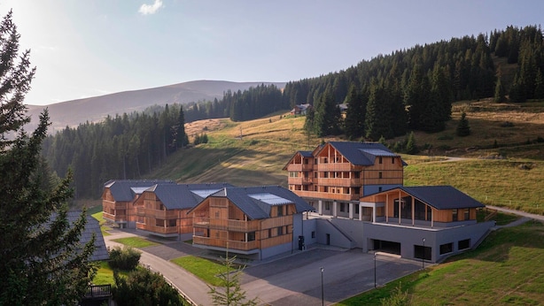 Un bâtiment moderne doté de nombreuses structures en bois et d’un grand bâtiment central à toit en pente, entouré d’un paysage vallonné et d’une forêt dense.