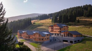A modern building with multiple wooden structures and a large central building with a sloped roof, surrounded by a hilly landscape and dense forest.