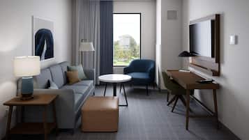 Une chambre d’hôtel moderne équipée d’un canapé, d’une table ronde, d’un fauteuil bleu, d’un bureau et d’un téléviseur fixé au mur.