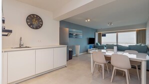 Une cuisine moderne avec des meubles blancs, un coin repas comprenant une table et des chaises blanches, une grande horloge au mur et une vue sur le salon, avec un canapé et un téléviseur.
