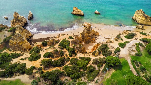 Vue aérienne d’une zone côtière caractérisée par des formations rocheuses, des eaux turquoise limpides et une plage de sable parsemée de végétation.
