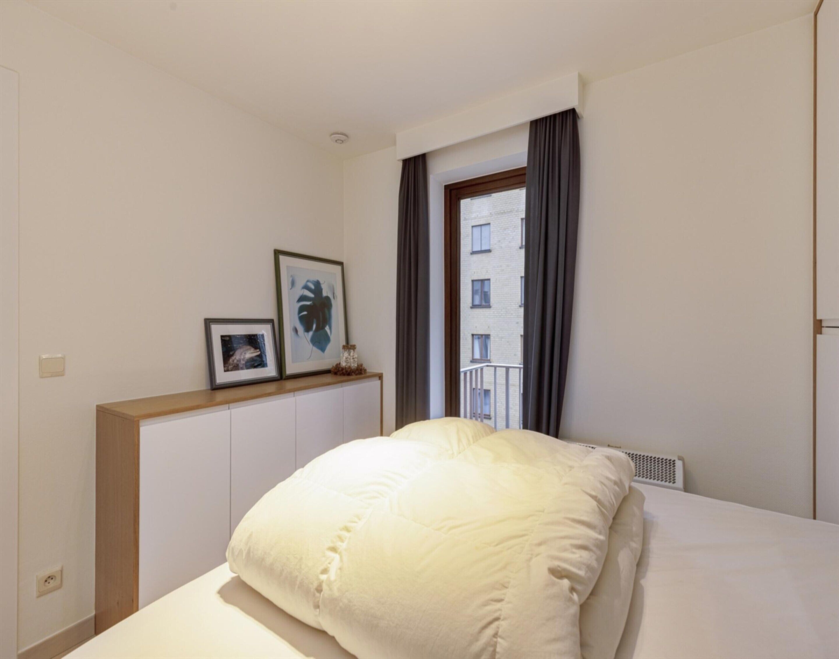 A hotel room with a bed, a window with curtains, a radiator, and framed pictures on the wall.