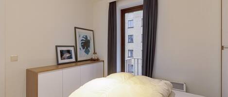 Une chambre d’hôtel avec un lit, une fenêtre avec des rideaux, un radiateur et des tableaux encadrés au mur.