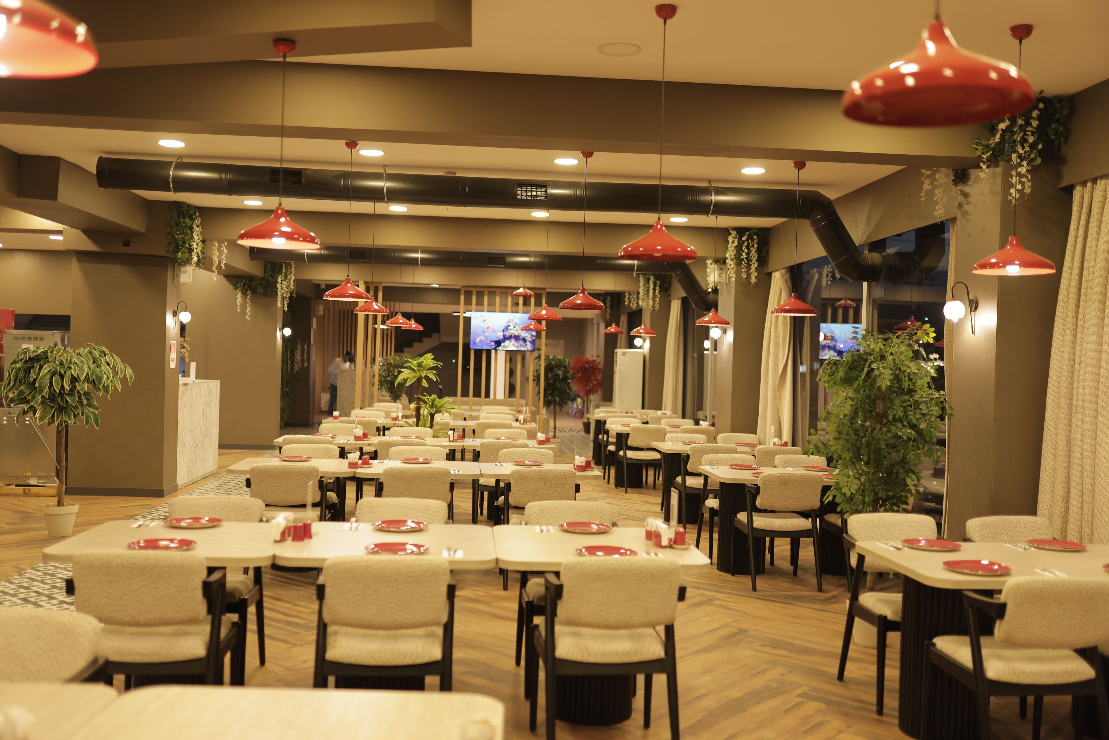 A spacious dining area with multiple tables and chairs, red pendant lighting, and potted plants.