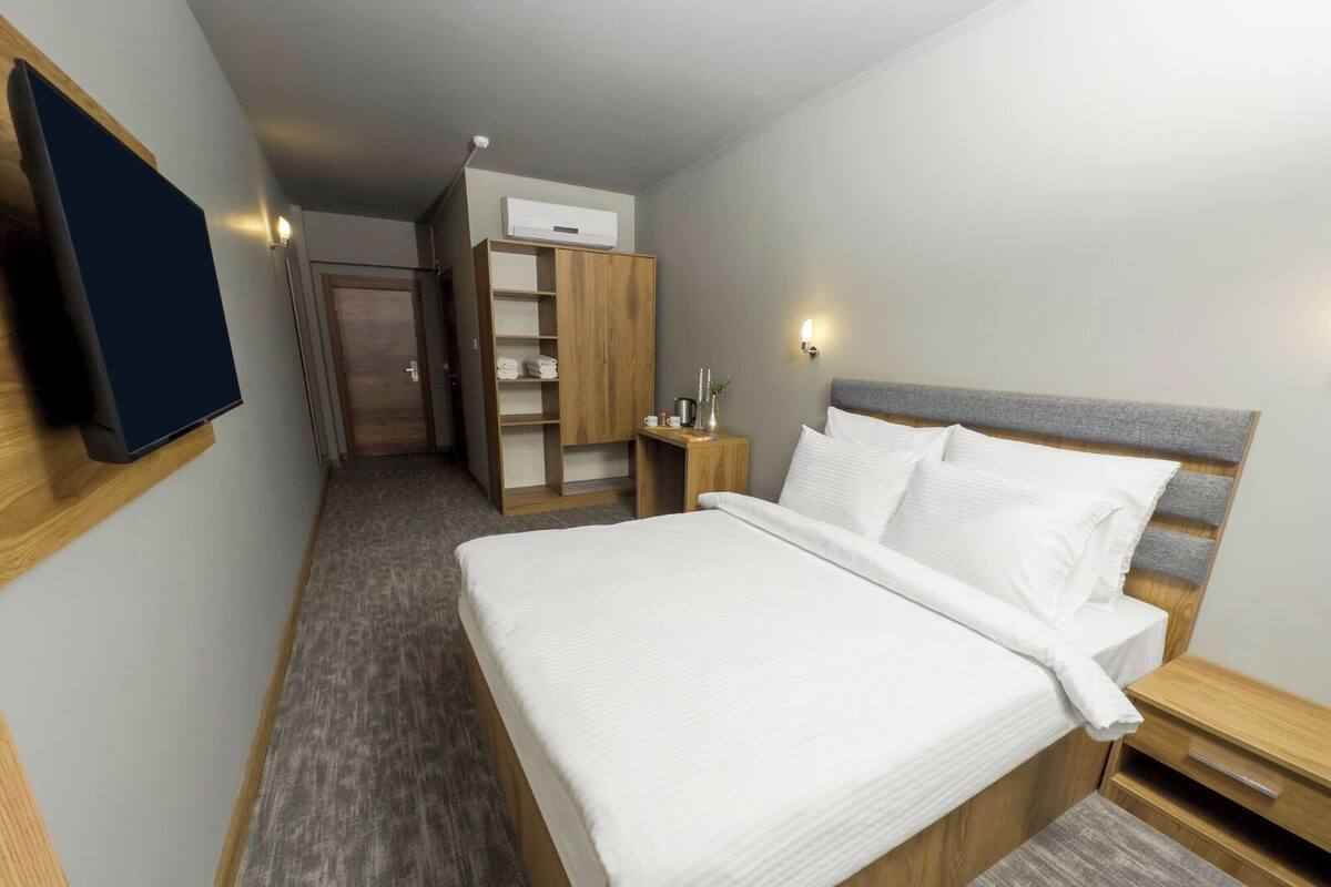 Quarto de hotel com uma cama grande, um guarda-roupa de madeira, uma pequena mesa de cabeceira e uma televisão de tela plana fixada na parede.