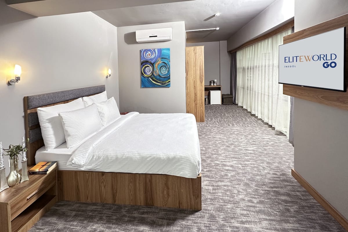 Quarto de hotel com uma cama grande, mesas de cabeceira, uma secretária pequena, uma cadeira e uma televisão mostrando o nome do hotel.