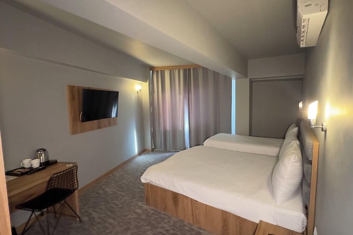 A hotel room with a bed, a desk with a chair, a television mounted on the wall, and a window with curtains.