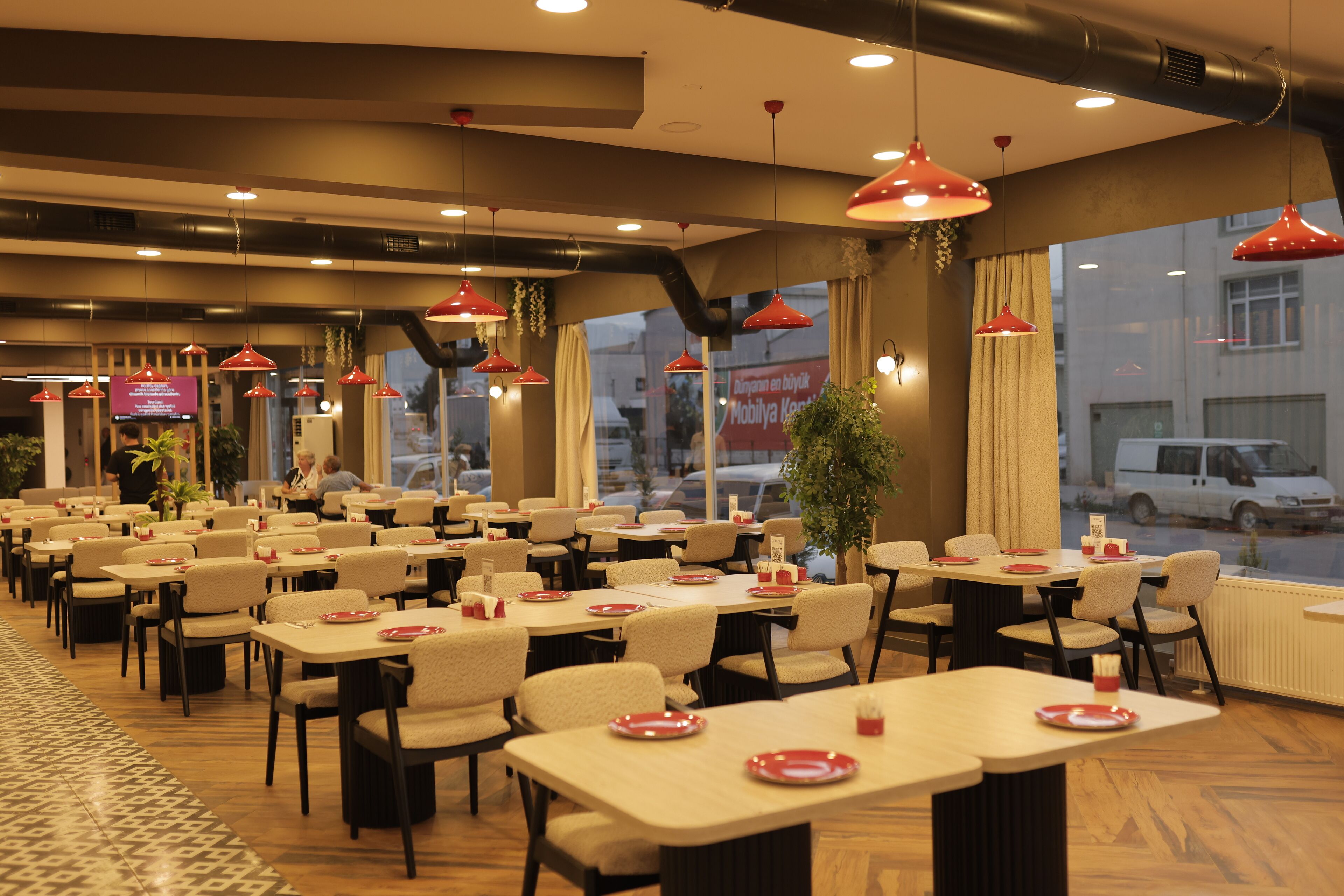 A restaurant with multiple tables and chairs, large windows, and hanging red pendant lights.