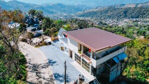 Una casa moderna de dos pisos amb un teulat vermell, envoltada de vegetació exuberant i turons.