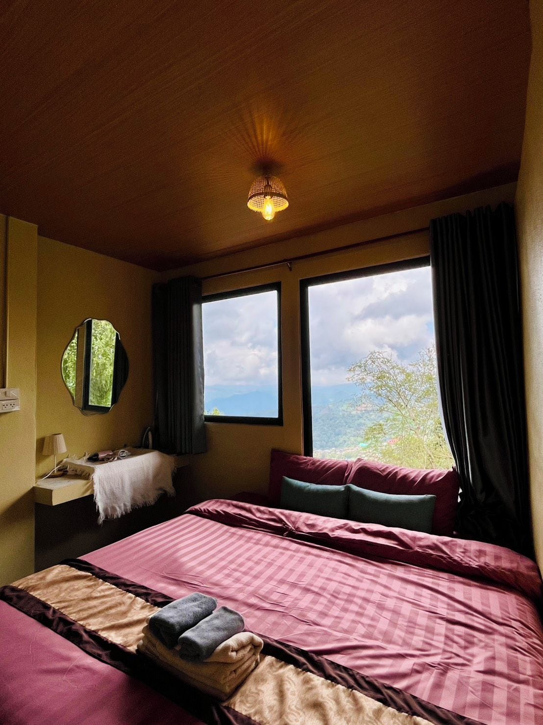 A room with a bed, a bedside table, a mirror, and a window with a view of the outdoors.