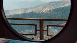 Una vista per una finestra amb un marc de fusta, sobre un paisatge ondulat amb una vila al peu.