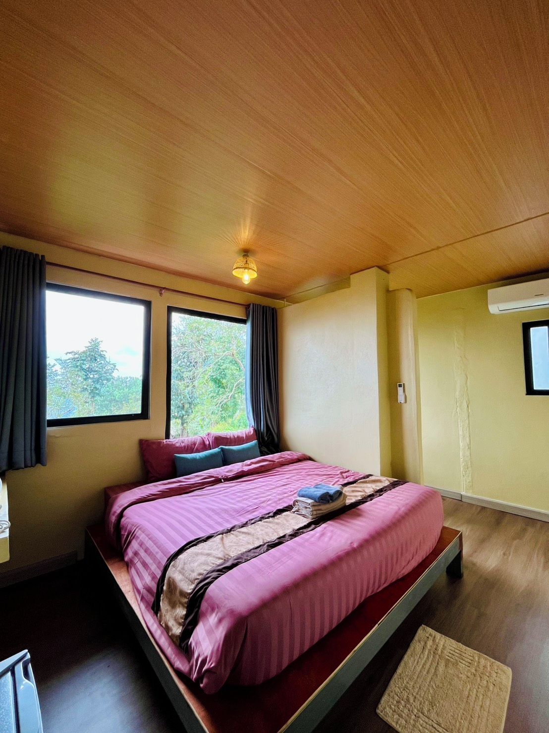A room with a bed, a window, an air conditioning unit, and a wooden ceiling.