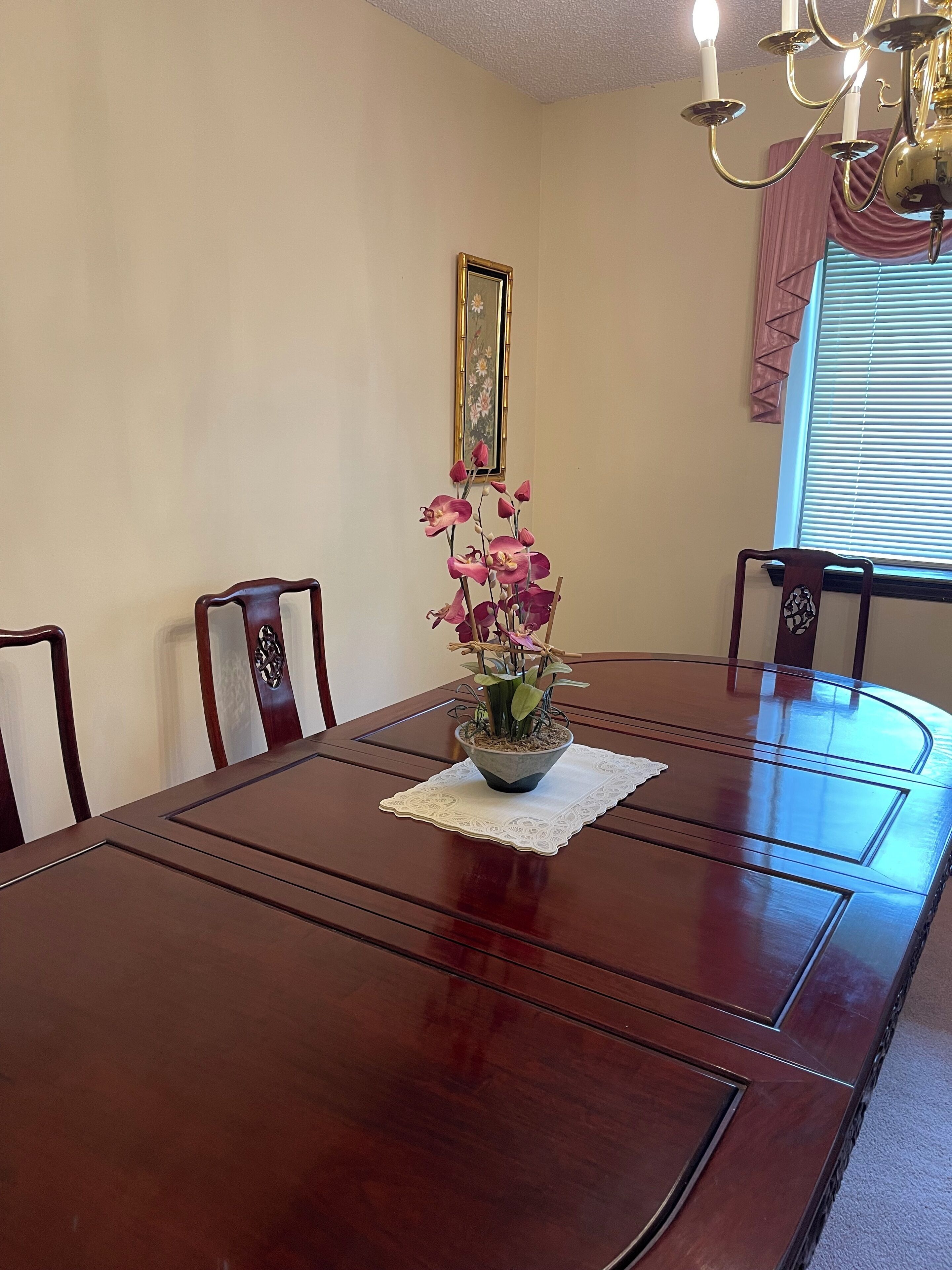 Une table à manger agrémentée d’un centre de table floral, entourée de chaises, dans une pièce dotée d’un lustre et d’une fenêtre avec des stores.